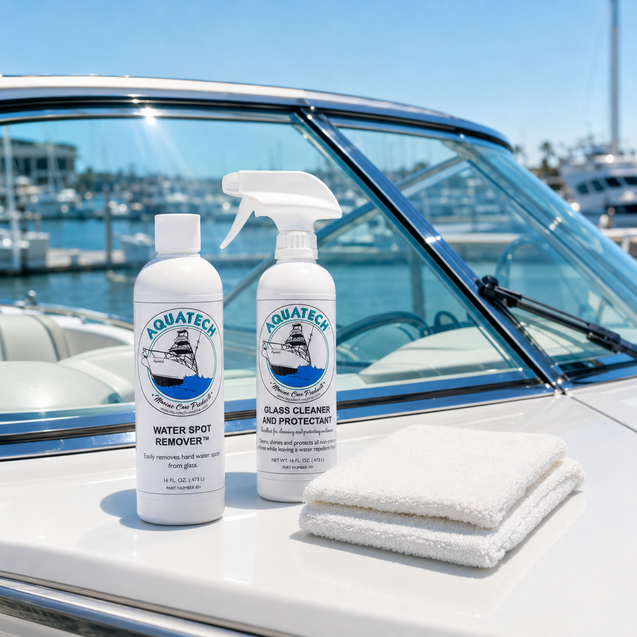 Two Aquatech cleaning products and a folded white microfiber cloth displayed on the white bow of a boat, with water and marina boats visible in the background.